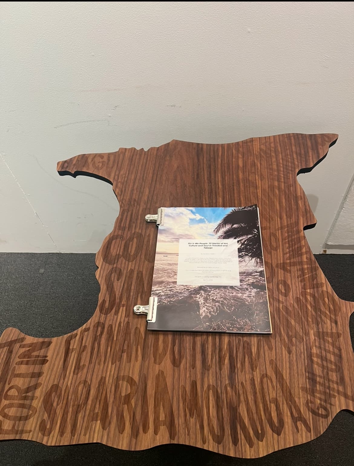 Custom Trinidad and Tobago shaped wooden table with 'Dis Is We People' engraved text and open magazine